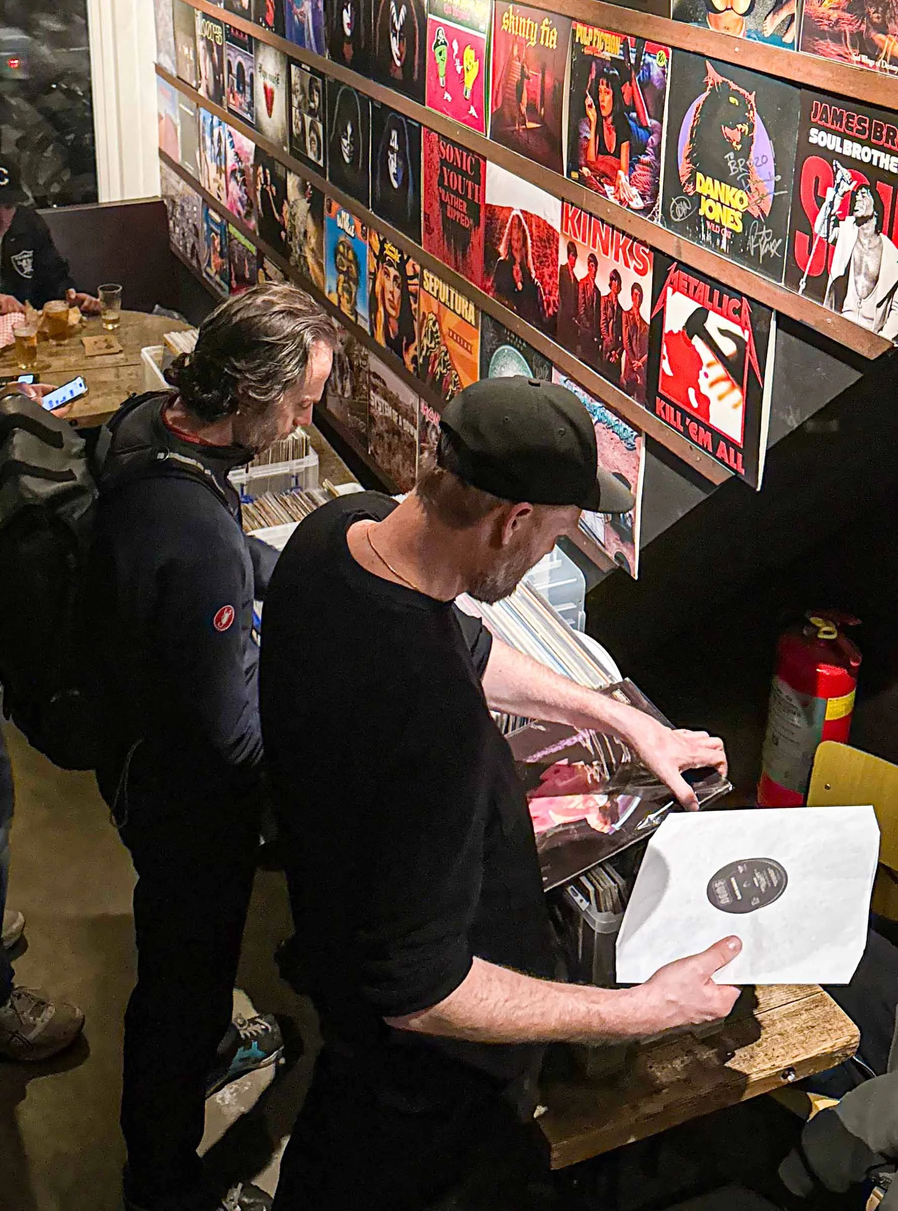 People browsing records at an Amsterdam Vinyl Club live event, with album covers on the wall above a wooden table.