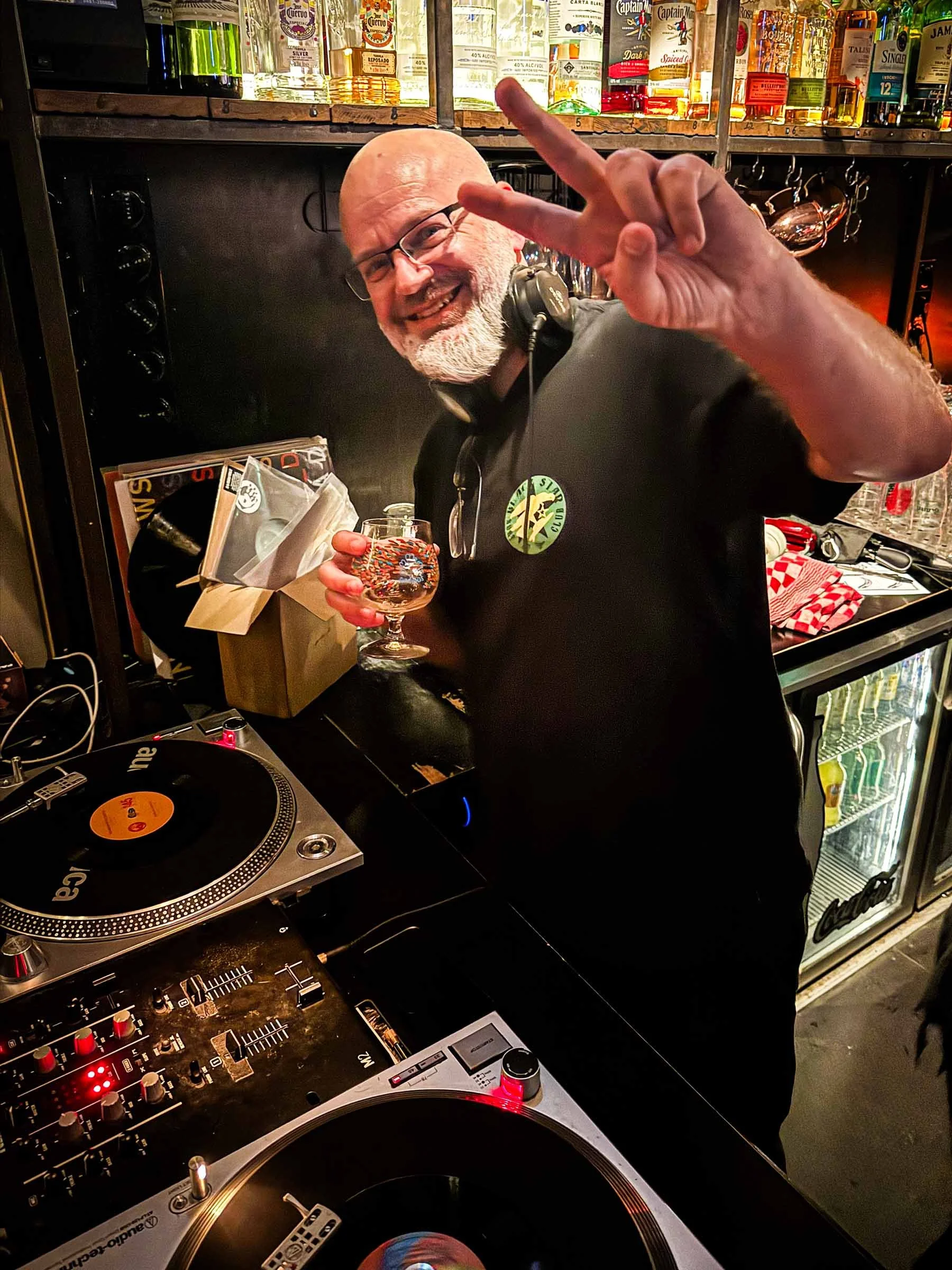 A man with a bald head and glasses, smiling and making a peace sign with his right hand, standing behind a DJ turntable setup in a bar or club. He is wearing a black shirt with a green circular logo. The background shows shelves with bottles of alcohol and glasses.
