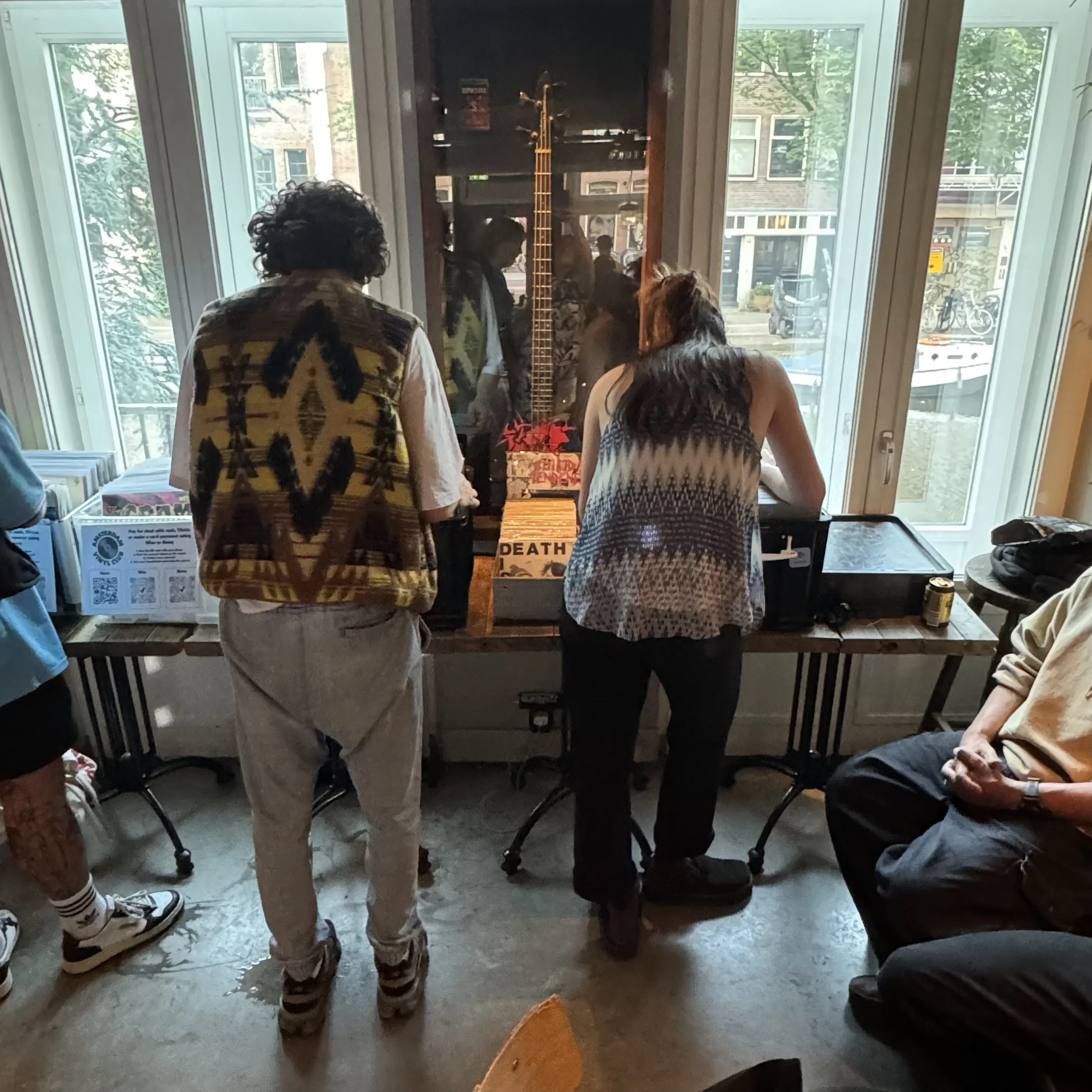People gathered around a table inside a room, looking at displayed items near a large window, with a guitar on the wall outside visible through the window.