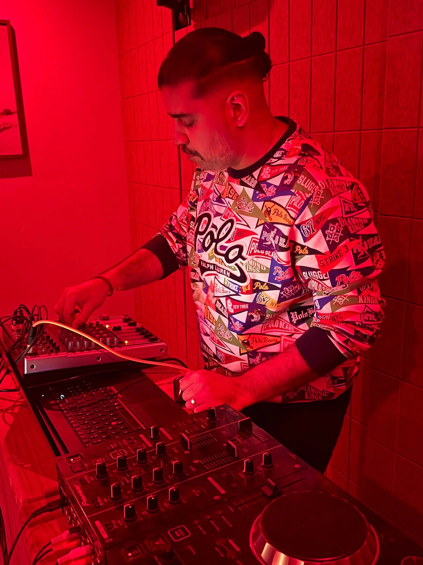 A man with dark hair and a mustache wearing a colorful patterned shirt is DJing with electronic music equipment. The scene is illuminated with red lighting and features a wooden wall behind him.