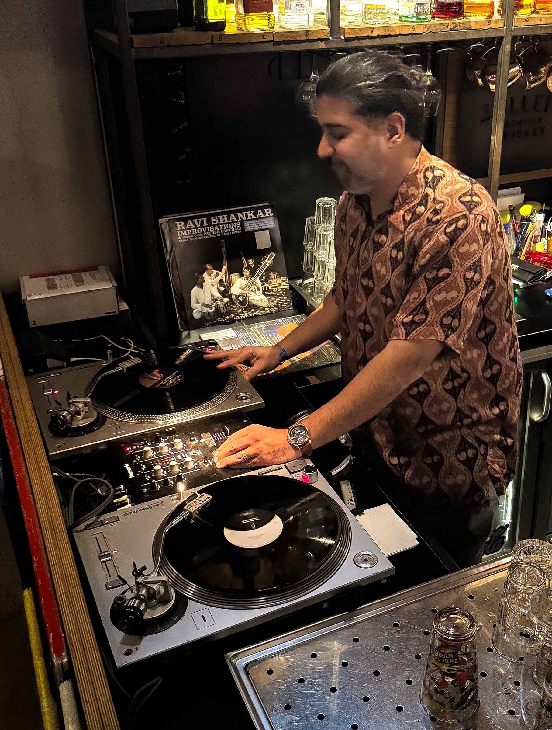 A DJ in a patterned shirt is mixing music with turntables and a mixer at a bar. There are shelves with bottles and glasses in the background, and some glass jars and bottles on the table in front.