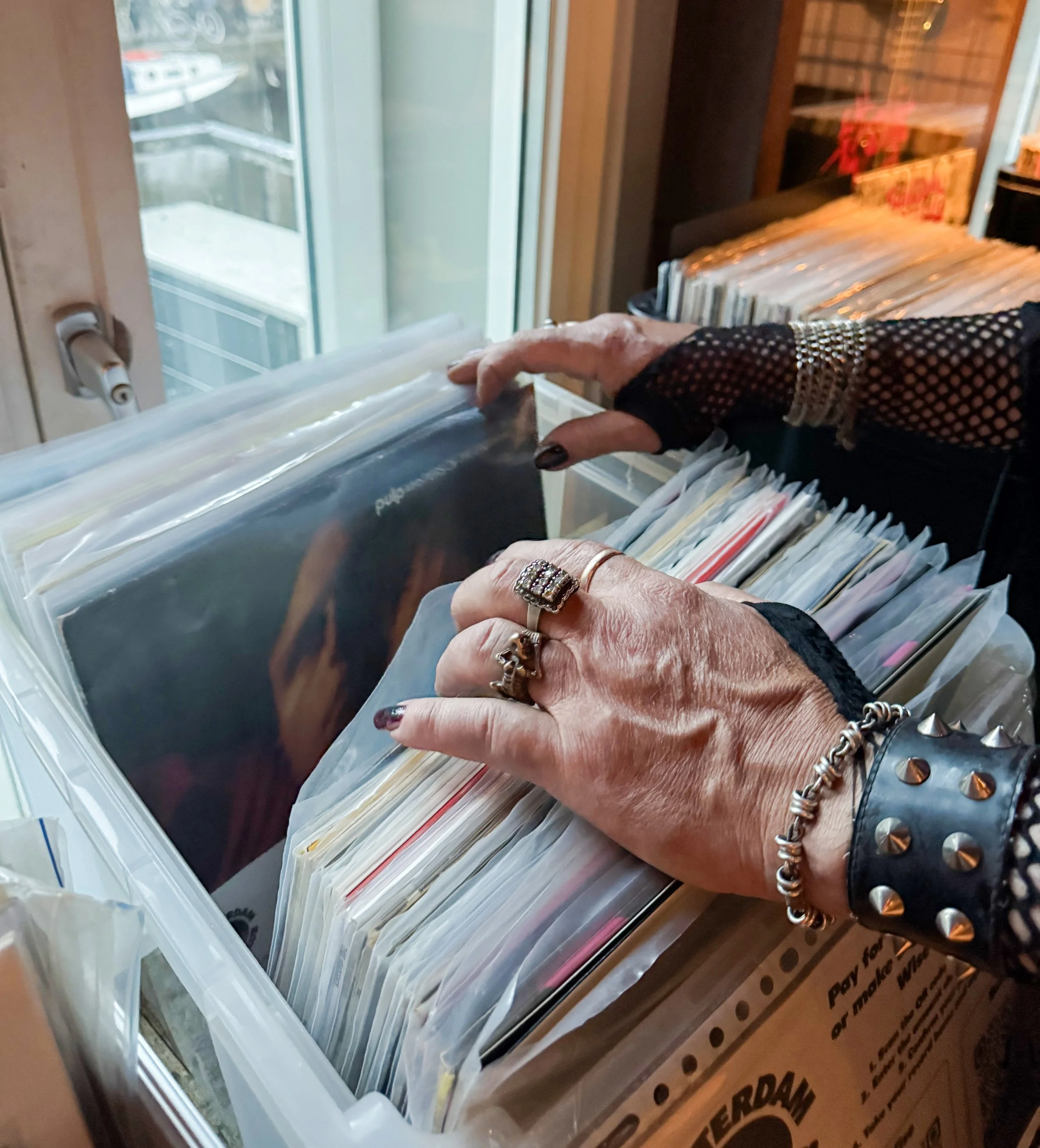 Close-up of a person's hands flipping through vinyl record covers in a cardboard box, with a window and shelves of records in the background.