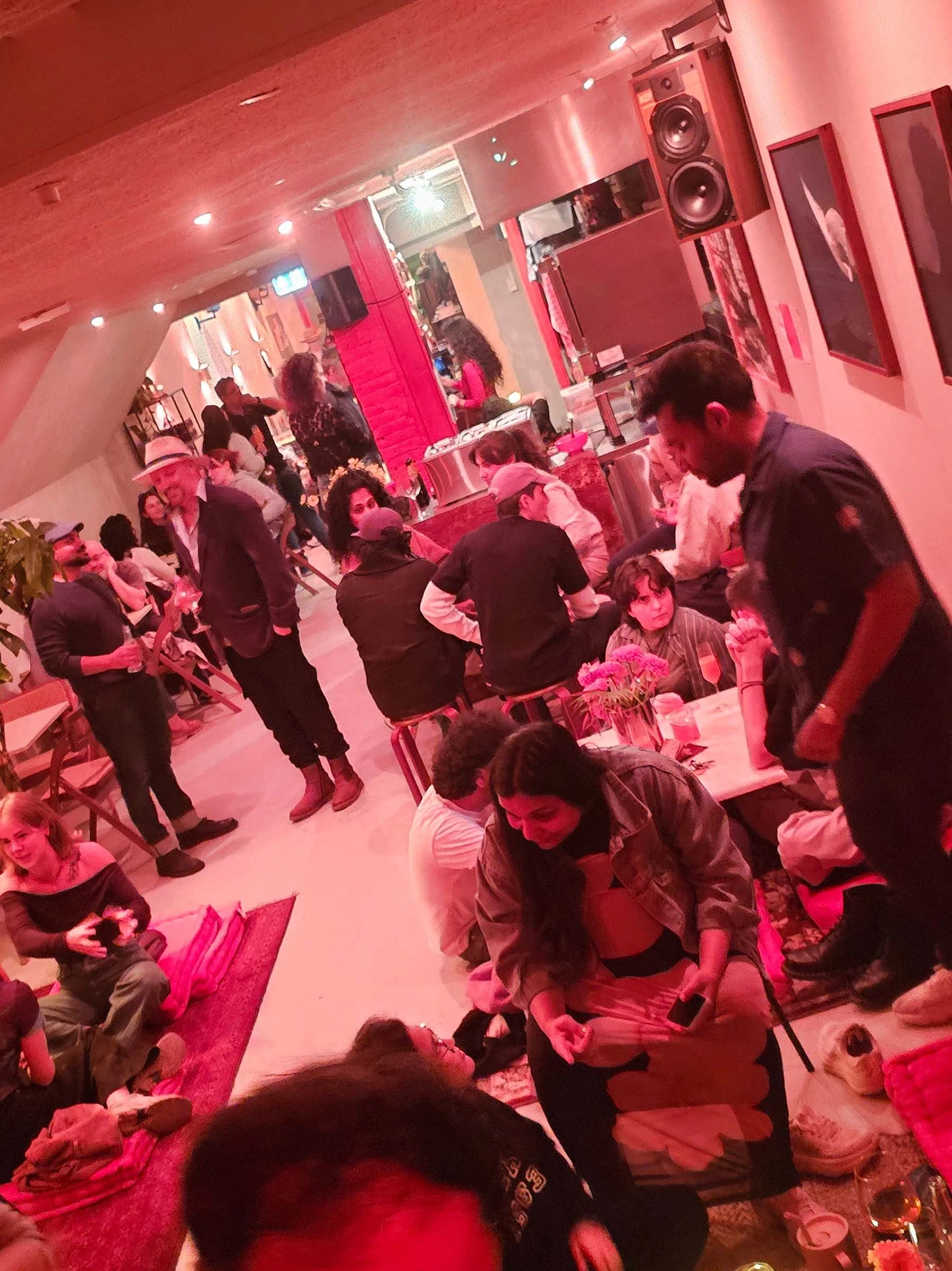 People socializing at Amsterdam Vinyl Club, an indoor space with warm lighting, some sitting on benches and others standing, with art on the walls and a large speaker.