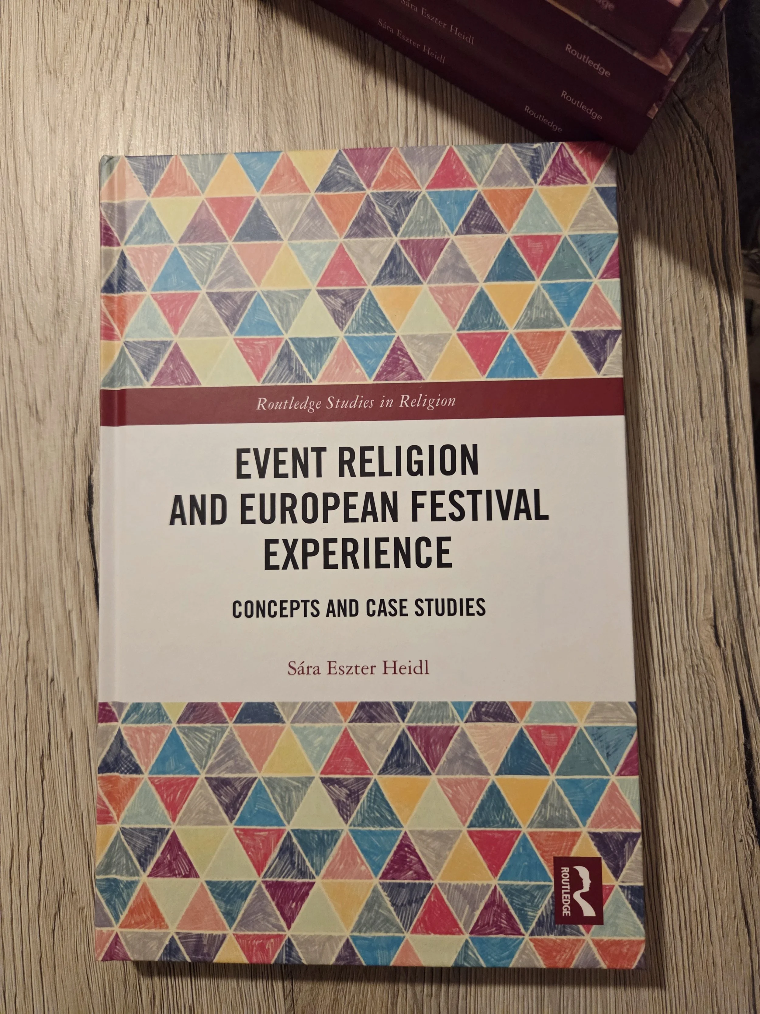 Photo of a book called "Event Religion and European Festival Experience. Concepts and Case Studies" by Sára Eszter Heidl, with colourful pattern with small triangles. On the top right corner a small part of bookspines of other copies of the same book