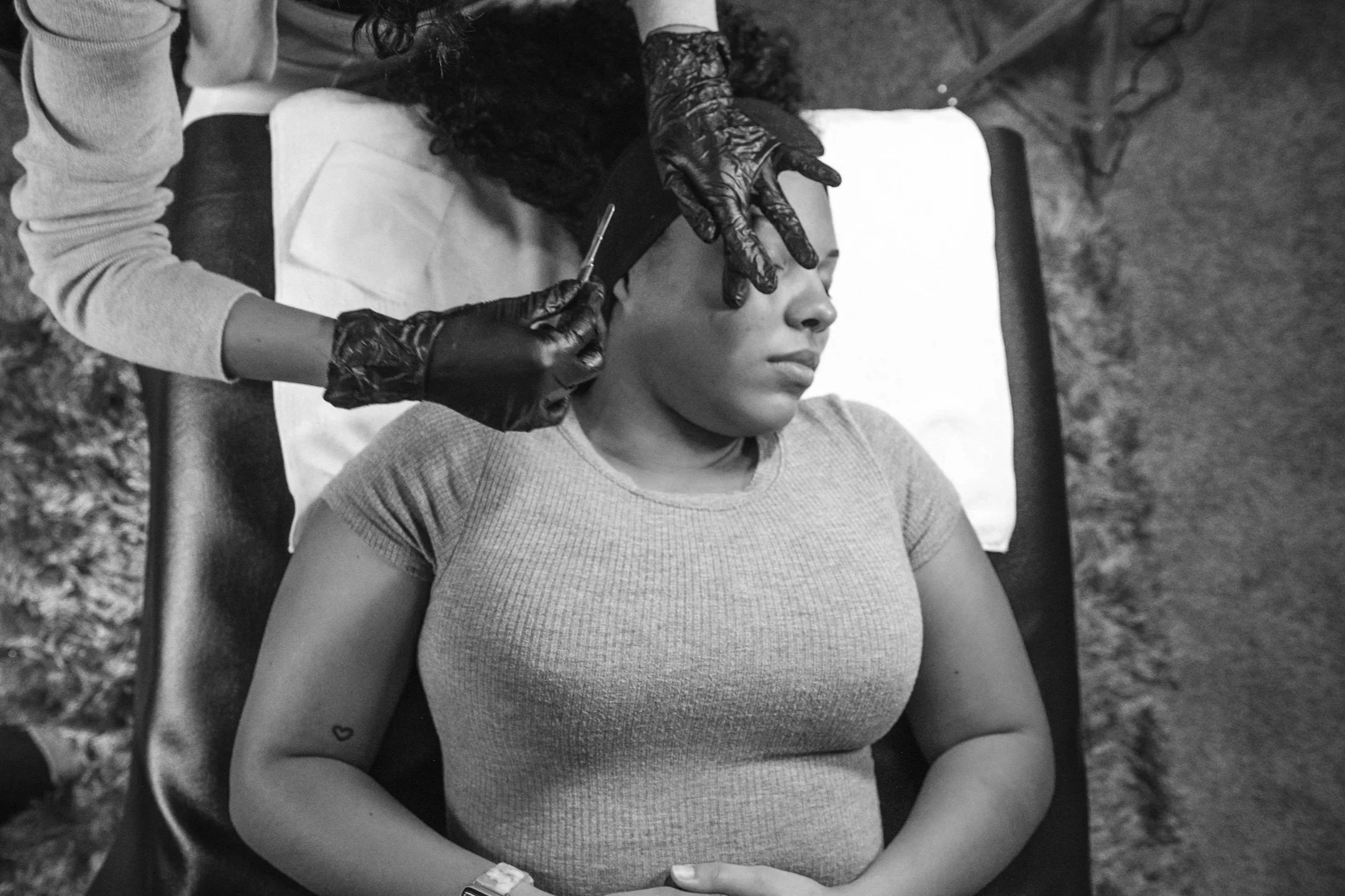 A woman lying on a table receiving a dermaplaning treatment on her face from a beautician wearing black gloves.