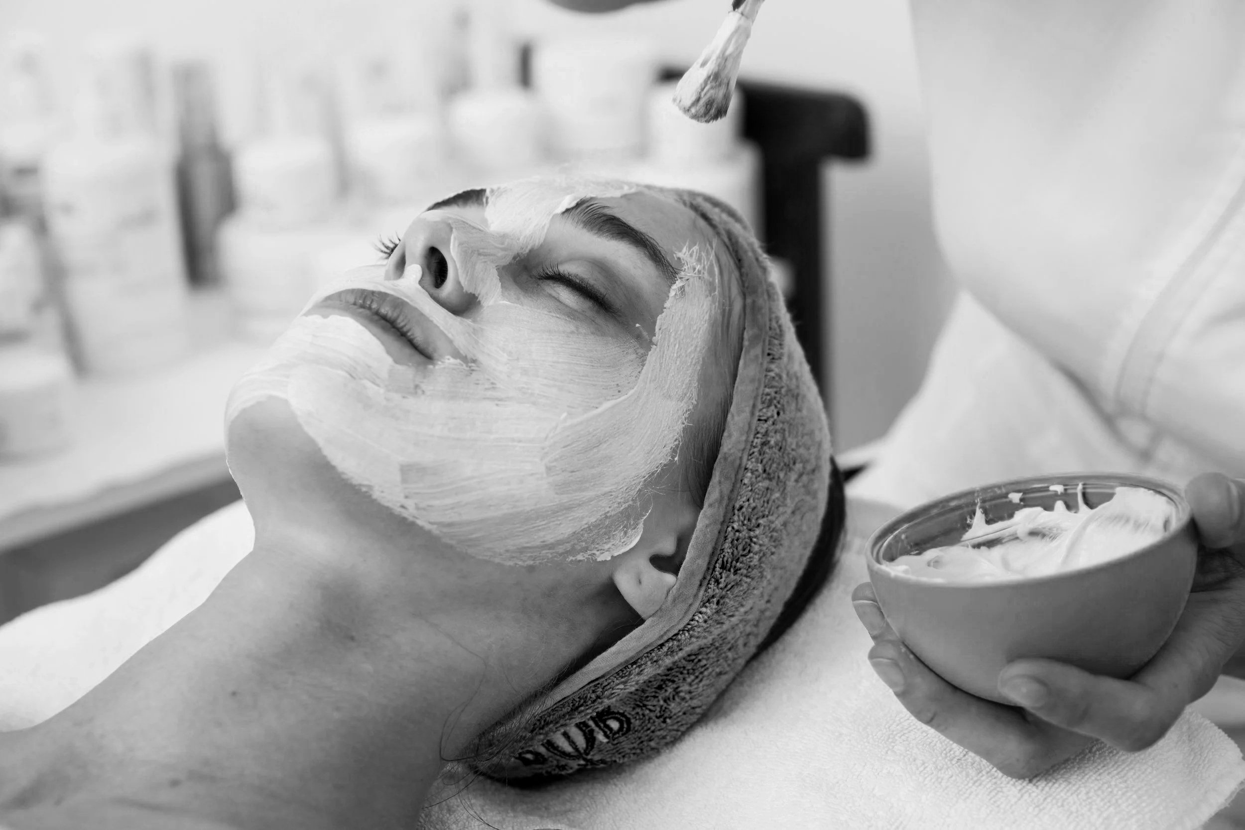 A woman receives a facial mask treatment at a spa, lying with her eyes closed, a towel wrapped around her head, as a skincare professional applies a white cream mask on her face.