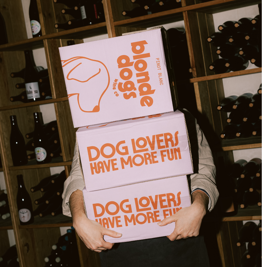 Person holding three pink boxes labeled "Dog Lovers Have More Fun" in front of a wine rack filled with various bottles.