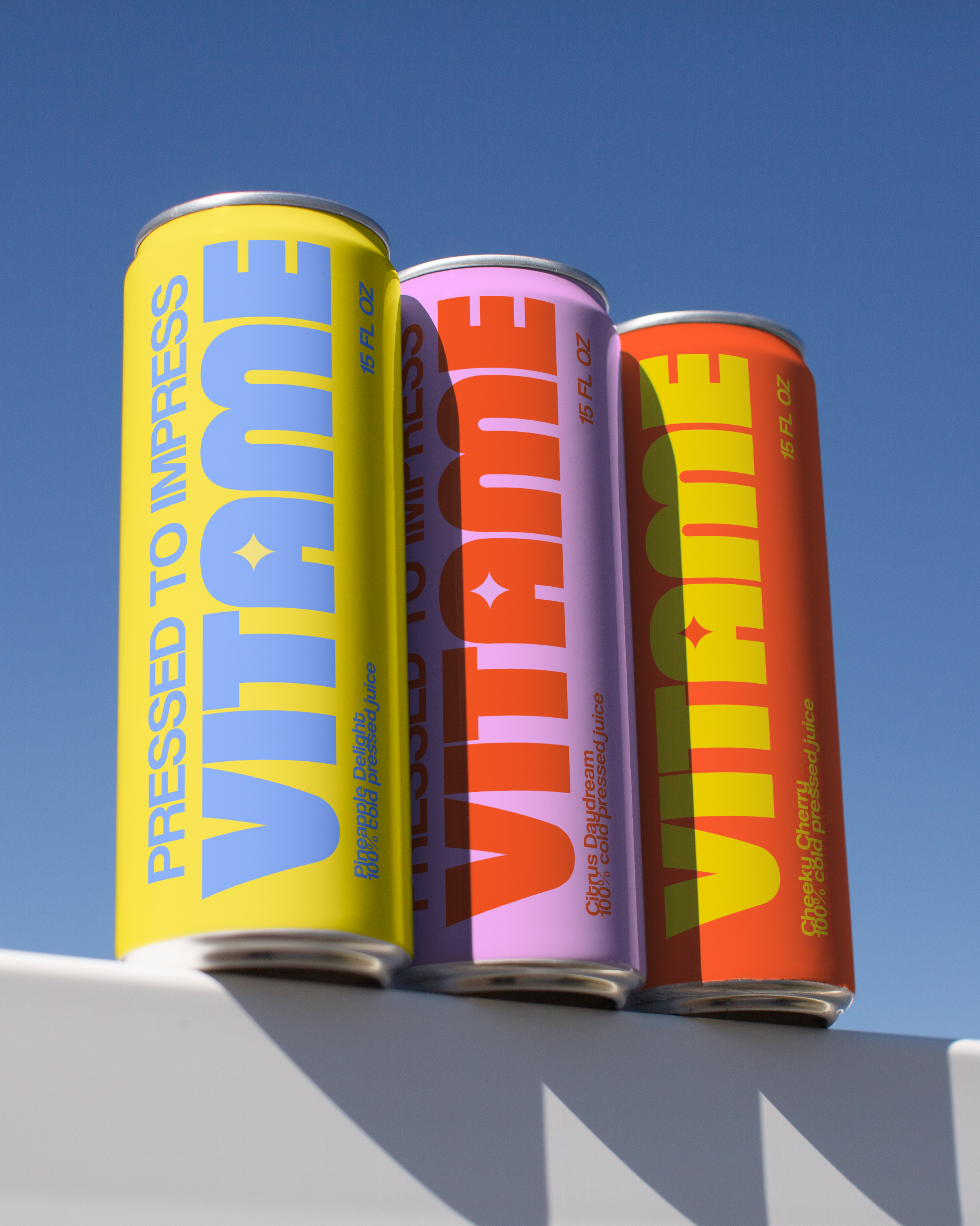Three cans of juice in yellow, pink, and orange colors, stacked outdoors against a bright blue sky.