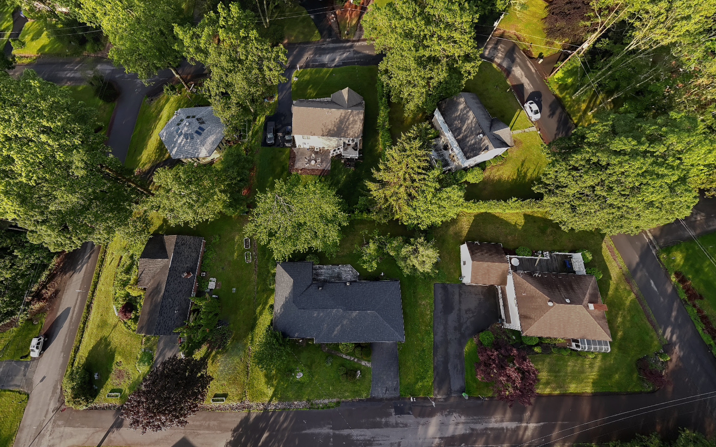 An aerial view of a residential neighborhood showing six houses, lush green trees, driveways, parked cars, and well-maintained lawns.