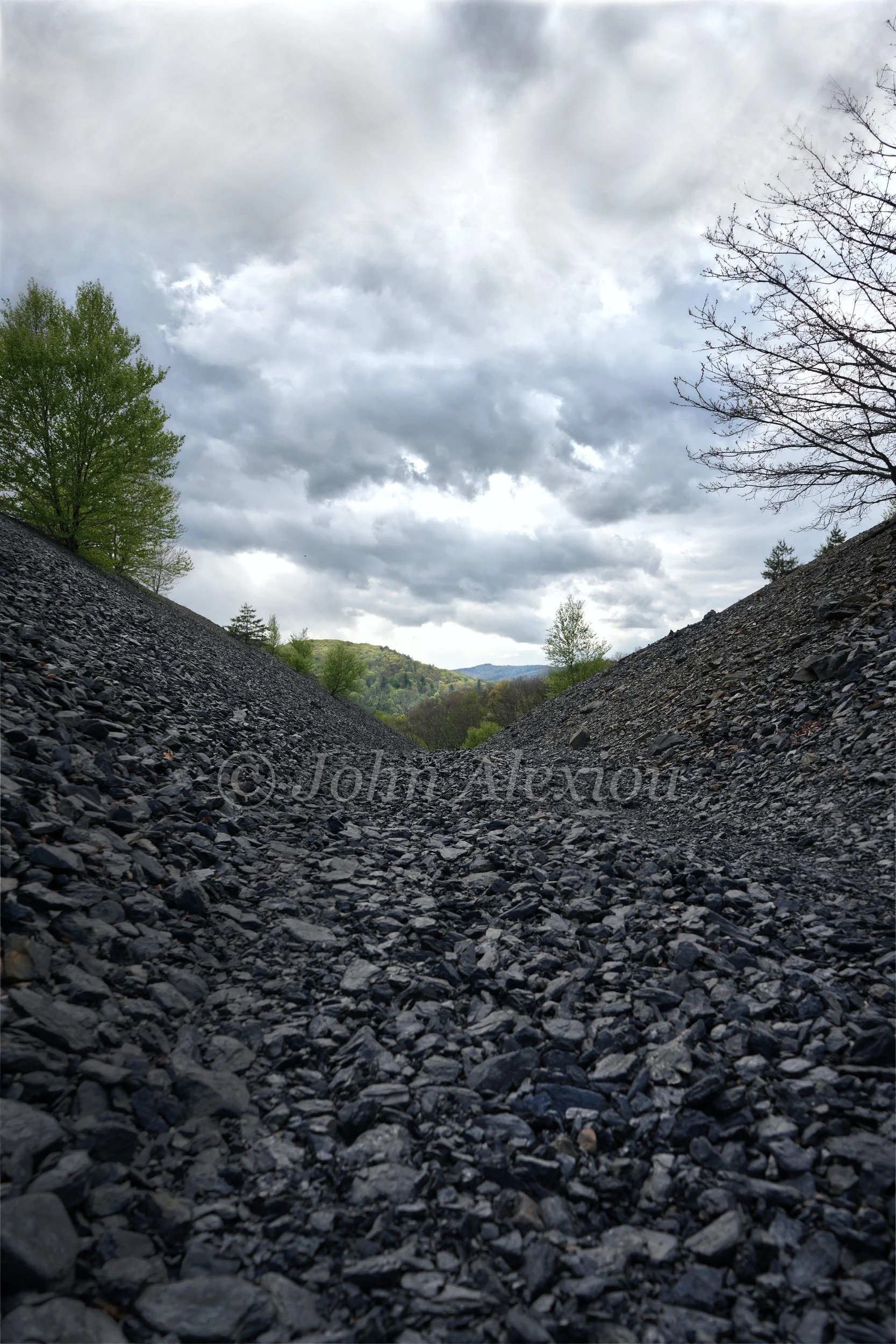 03 · A Break in the Clouds: A rocky passage opens toward trees beneath a dramatic cloud-filled sky.

© John Alexiou · 2025 · Mocanaqua
