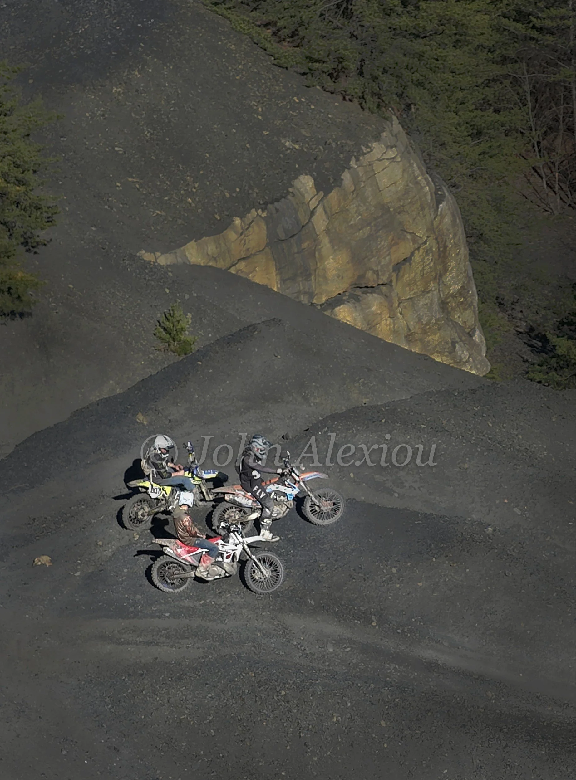 08 · Passing Through: Motorcycle riders pause beneath an exposed rock face on the dark mining terrain.

© John Alexiou · 2025 · Mocanaqua
