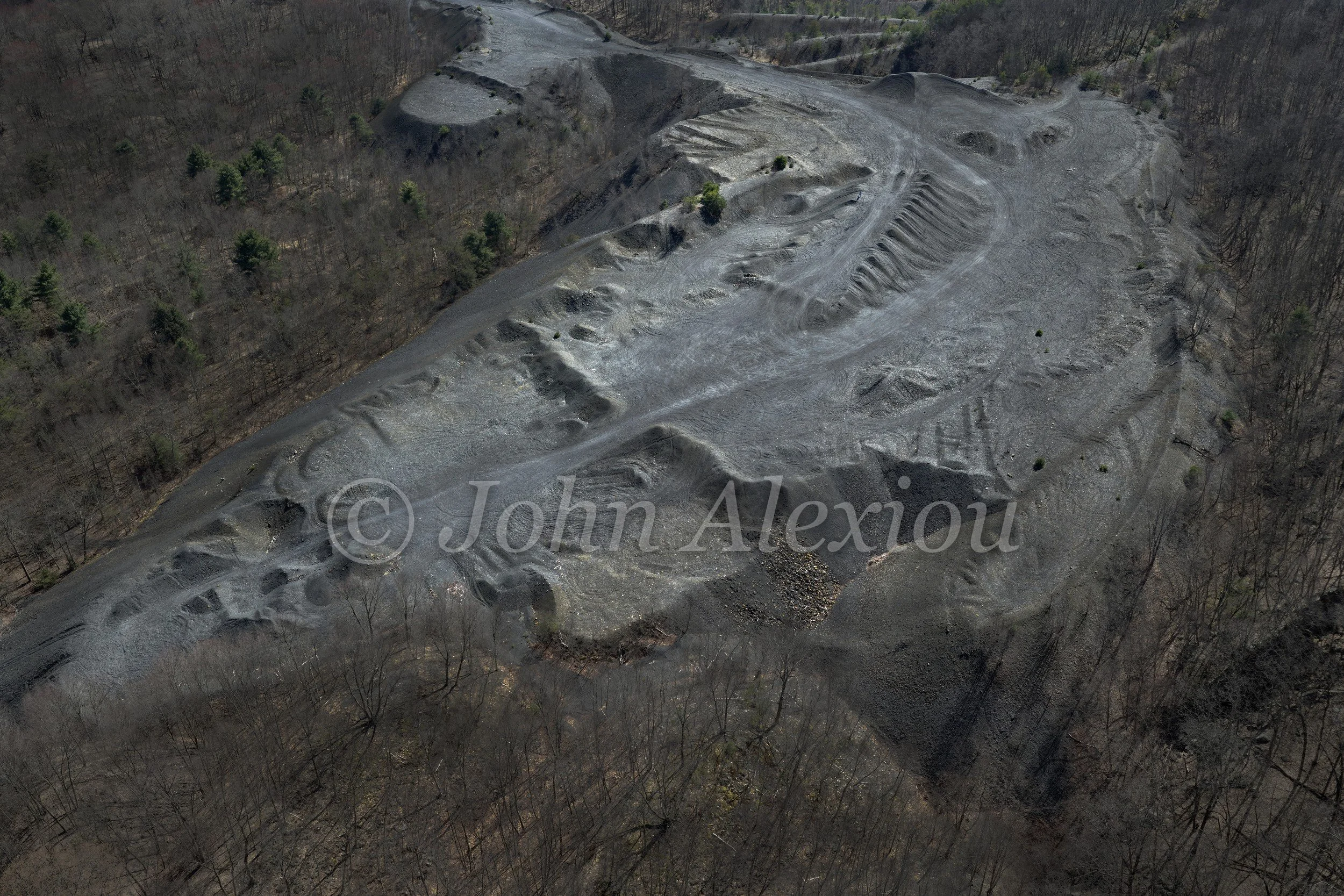 05 · The Shape of What Remains: An aerial view reveals worn slopes, tracks and dark folds among woodland.

© John Alexiou · 2025 · Mocanaqua