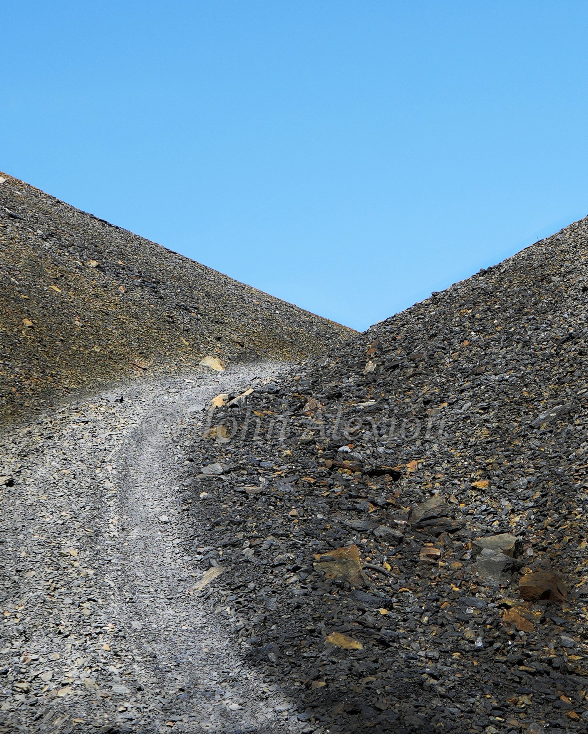 02 · Between the Banks: A narrow track climbs between two dark banks beneath a clear blue sky.

© John Alexiou · 2025 · Mocanaqua