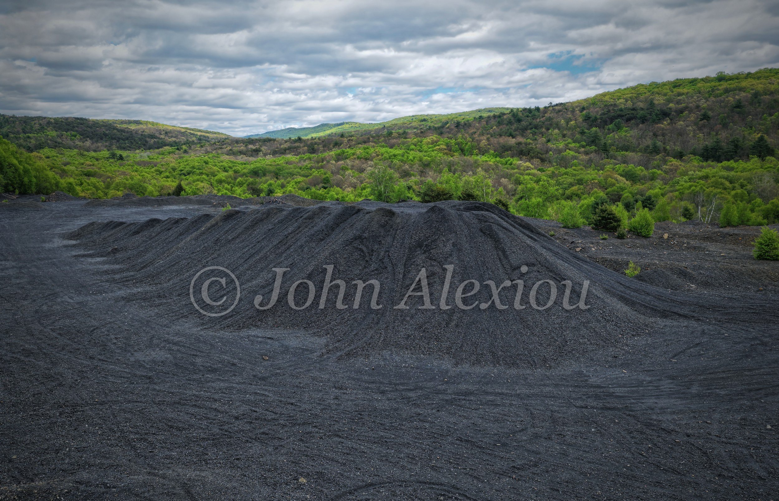 01 · The Land Remembers: Dark mining waste banks beneath a green wooded ridge and cloudy sky.

© John Alexiou · 2025 · Mocanaqua