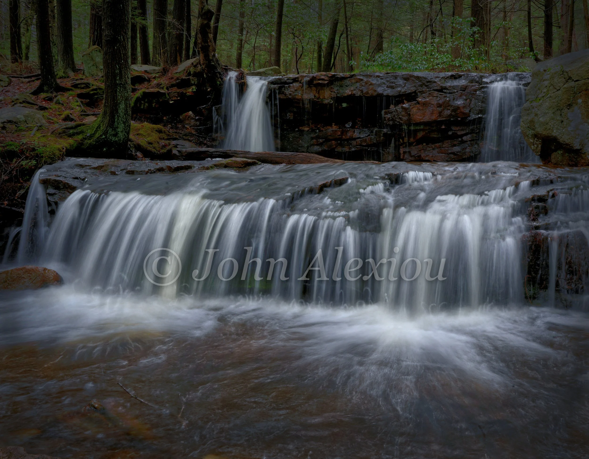 06 · A Quiet Place to Stay
Two small falls feed a wide curtain of water, framed by tree trunks and moss along the bank.
© John Alexiou · 2026 · Northeast Pennsylvania