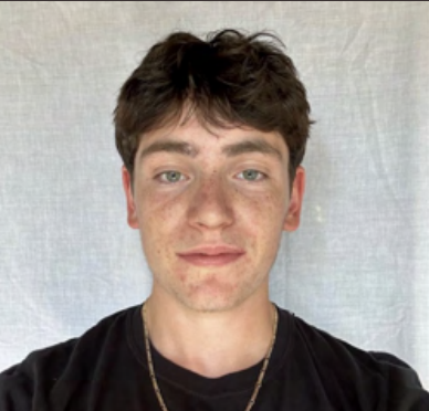 A young man with brown hair and blue eyes, wearing a black shirt and a gold chain, standing in front of a neutral background.
