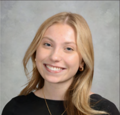 Young woman with blonde hair smiling, wearing a black top and gold earrings, against a gray background.