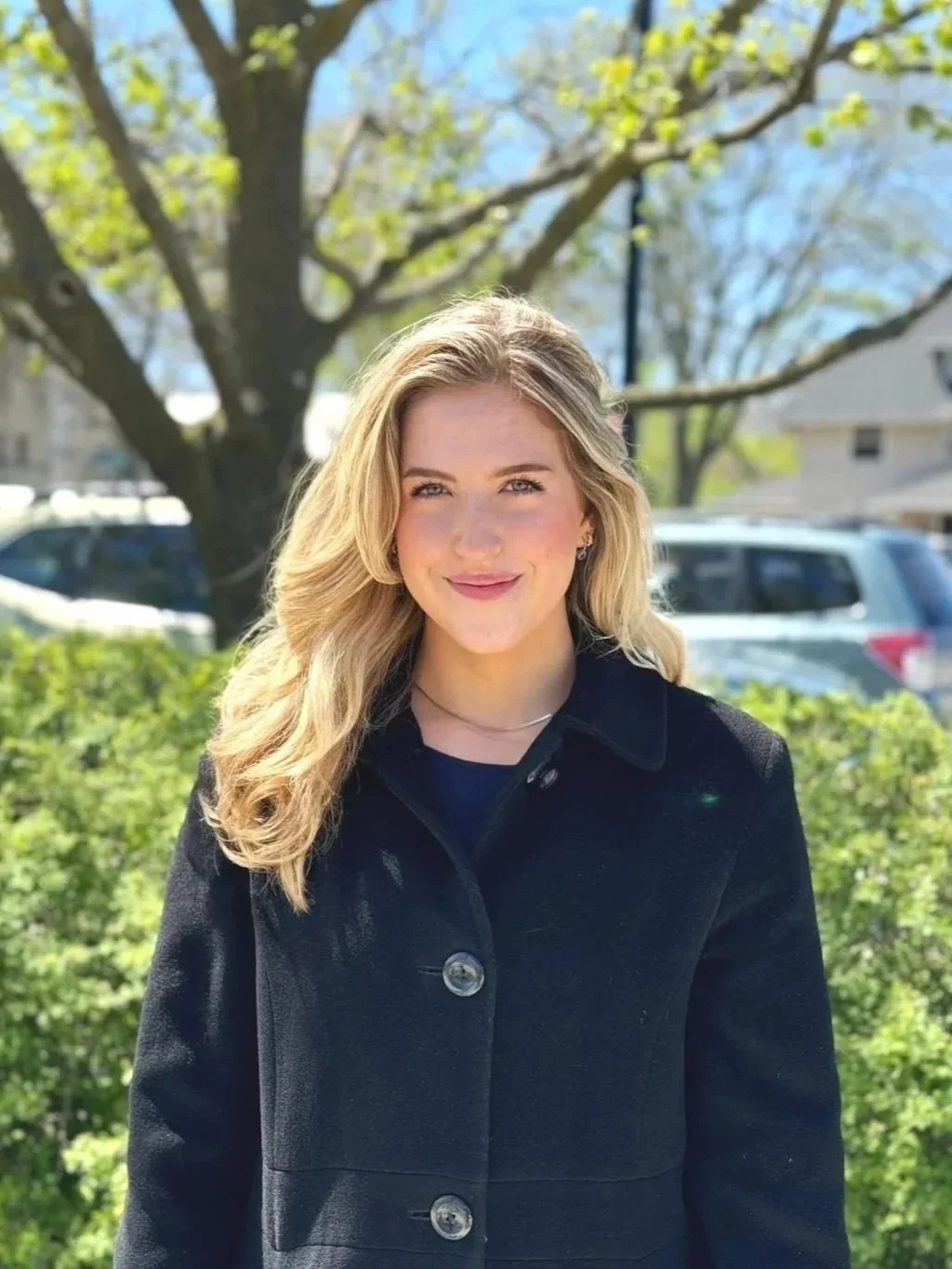 A young woman with long blonde hair smiling outdoors on a sunny day, standing in front of a tree and greenery, with parked cars and houses in the background.