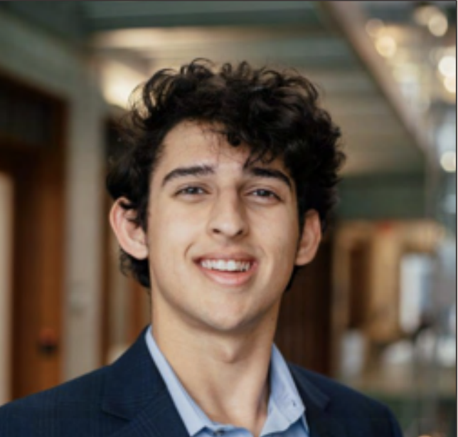 A young man with curly dark hair, wearing a navy blazer and a light blue shirt, smiling in an indoor setting with blurred background.