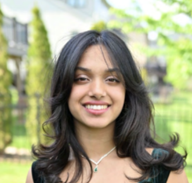 A young woman with black hair, smiling outdoors with trees and a fence in the background.