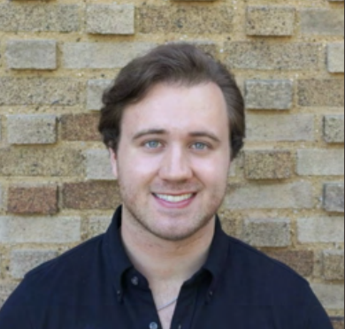 A young man with light skin, brown hair, and blue eyes smiling in front of a brick wall.