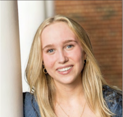 A young woman with blond hair and blue eyes smiling and leaning against a wall.