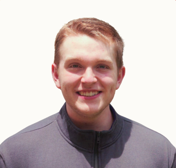 Portrait of a young man with short red hair, smiling, wearing a gray zip-up jacket, standing against a plain white background.