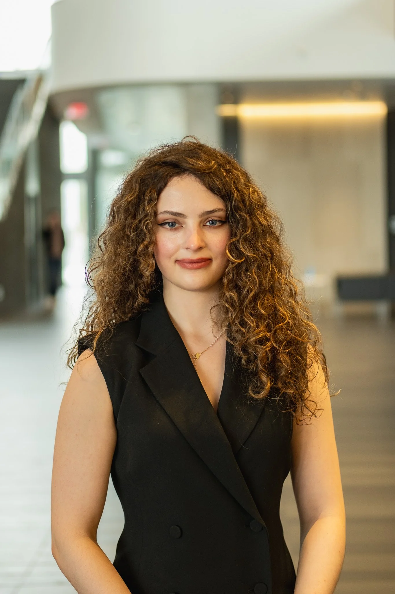 A woman with curly brown hair, wearing a sleeveless black blazer, standing in a modern indoor space with blurred background.
