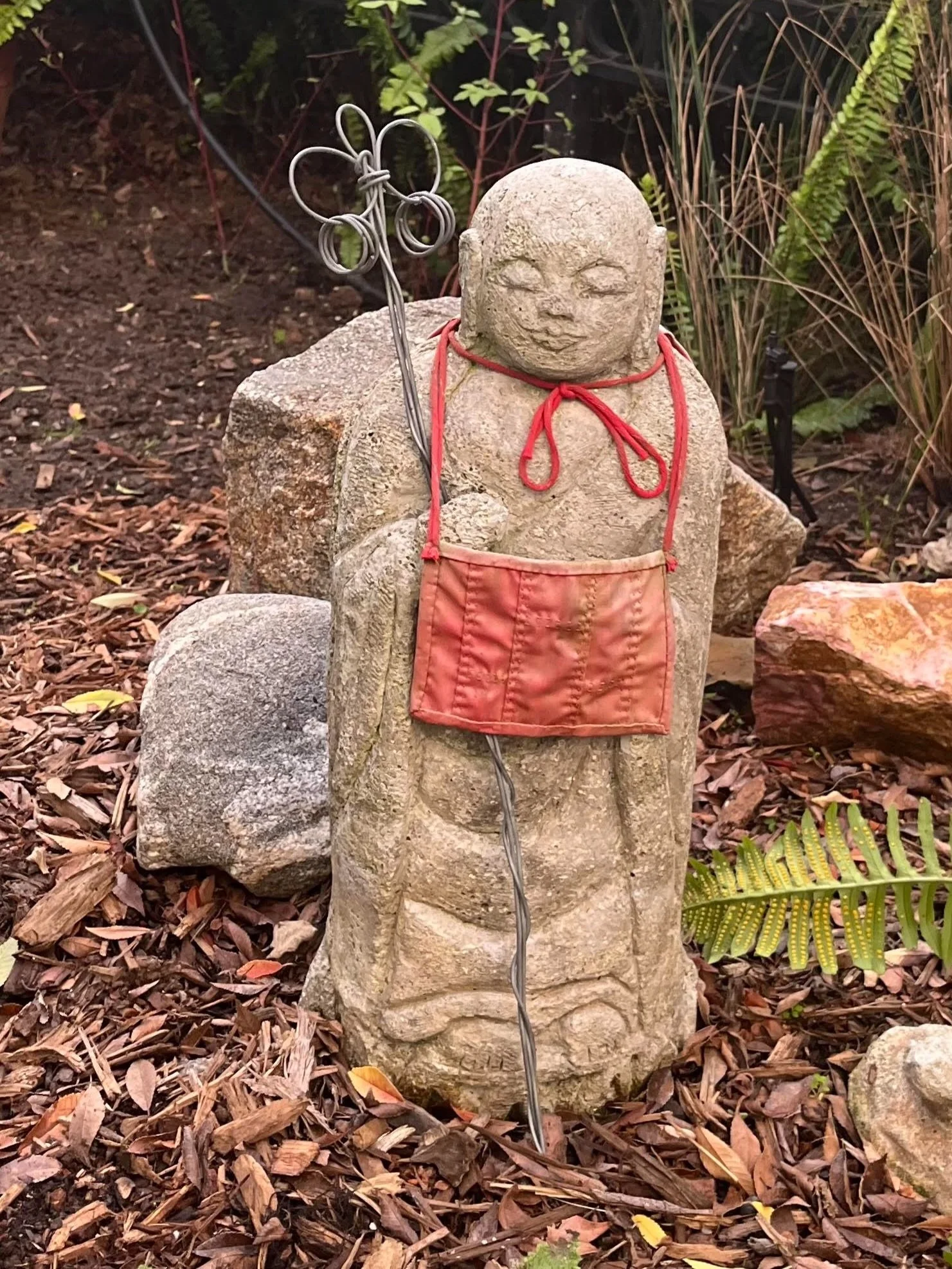 Garden sculpture of a small, smiling, carved stone figure with closed eyes, wearing a small orange rakusu, holding a metal rod with a decorative top, surrounded by soil and plants.