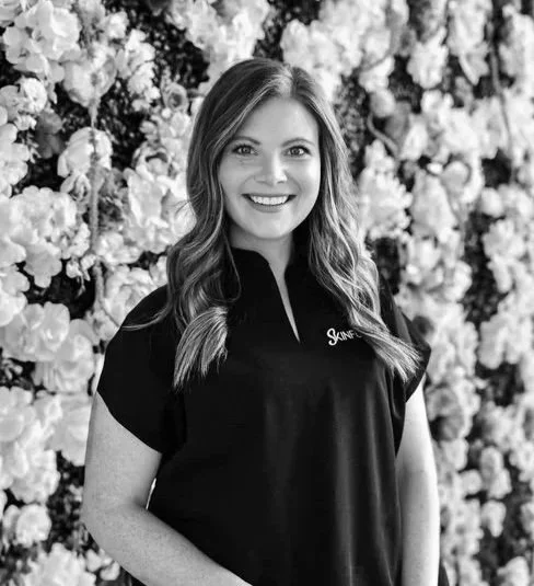 A woman with long hair smiling, standing in front of a floral backdrop, wearing a dark shirt.