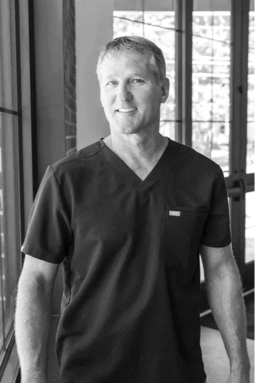 A smiling man in black medical scrubs standing indoors near large windows.
