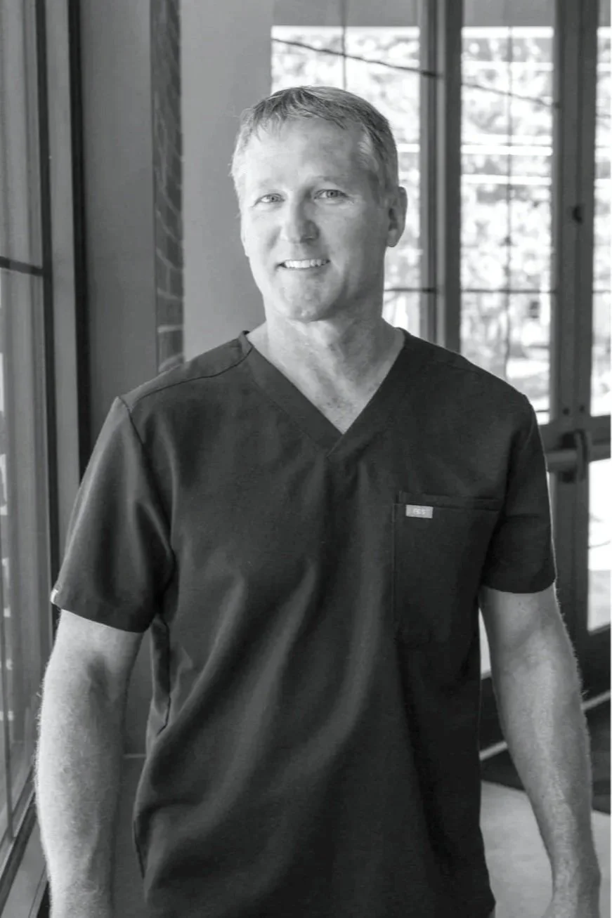 A smiling man in black medical scrubs standing indoors near large windows.