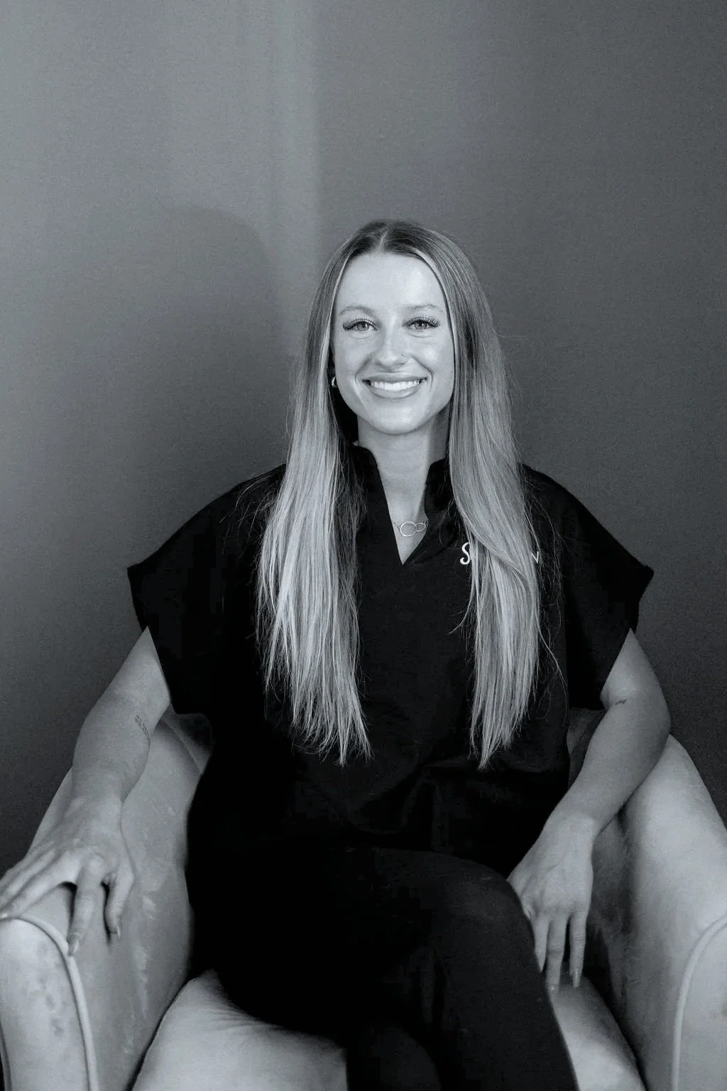 A woman with long straight hair smiling, sitting on a plush armchair, wearing a black shirt, in front of a plain wall.