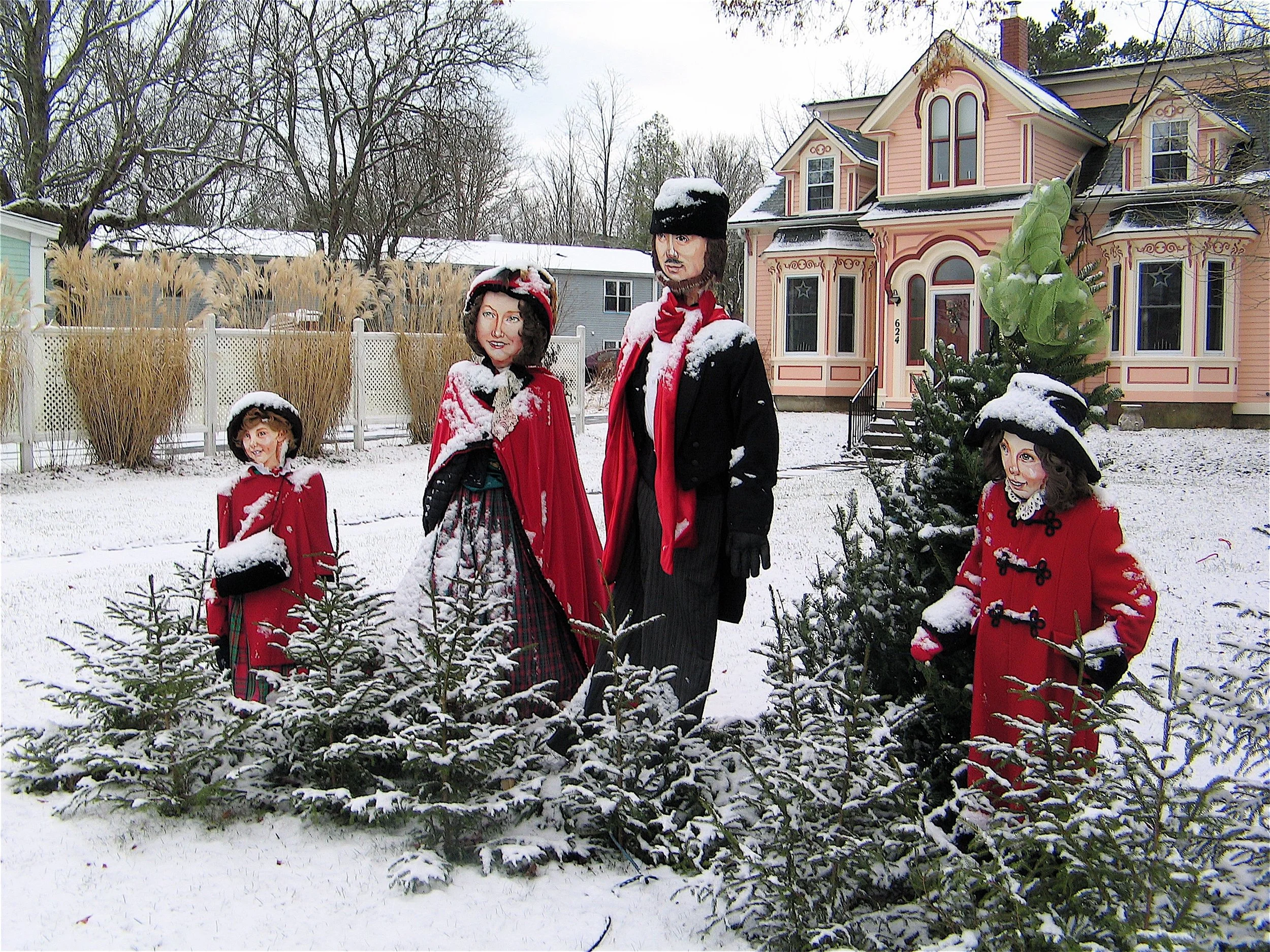 Snow-covered yard featuring four mannequin figures dressed in Victorian-style clothing, a decorated Christmas tree, and a large Victorian-style house in the background.