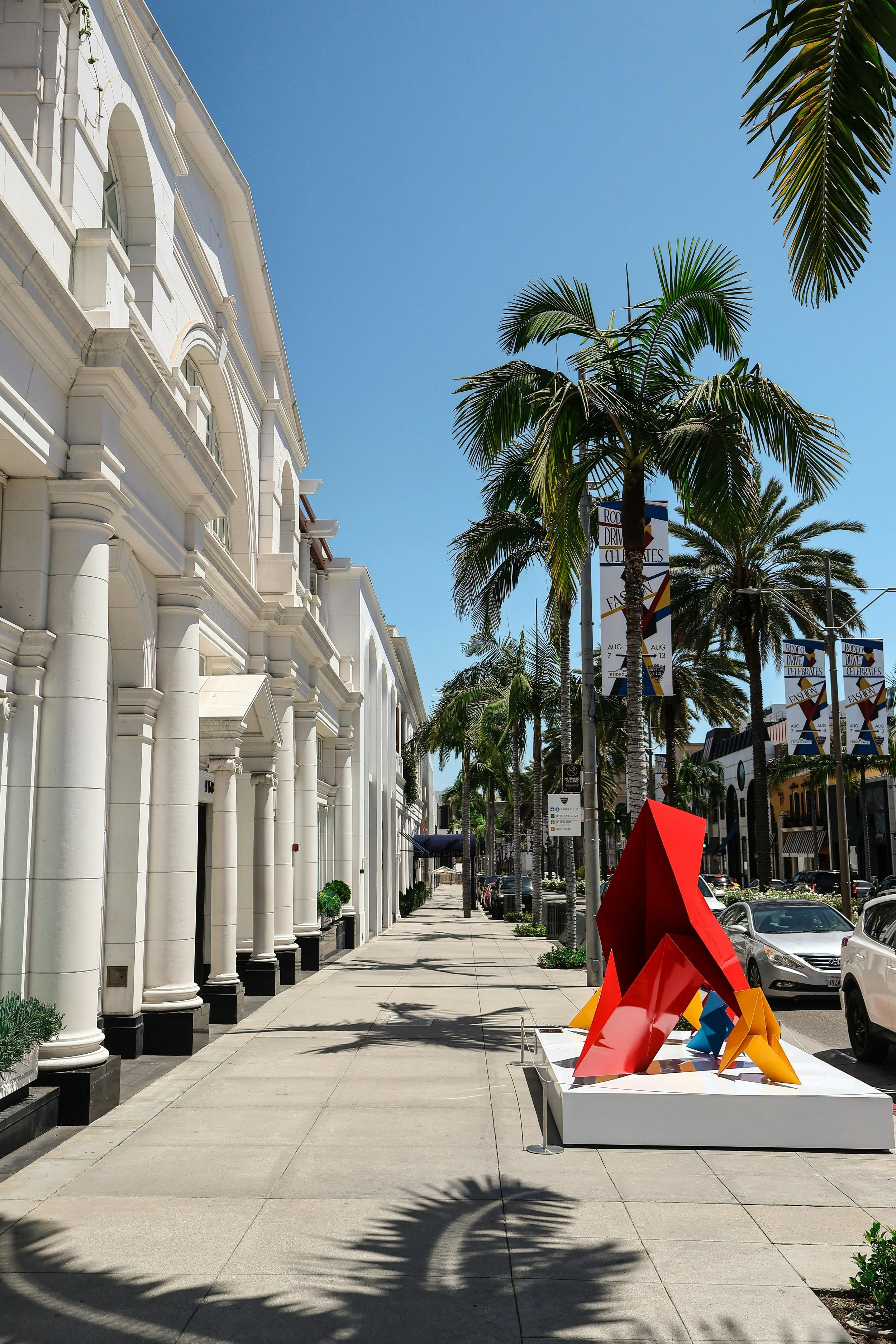 City street with white buildings, palm trees, and colorful modern sculpture on sidewalk during sunny day.