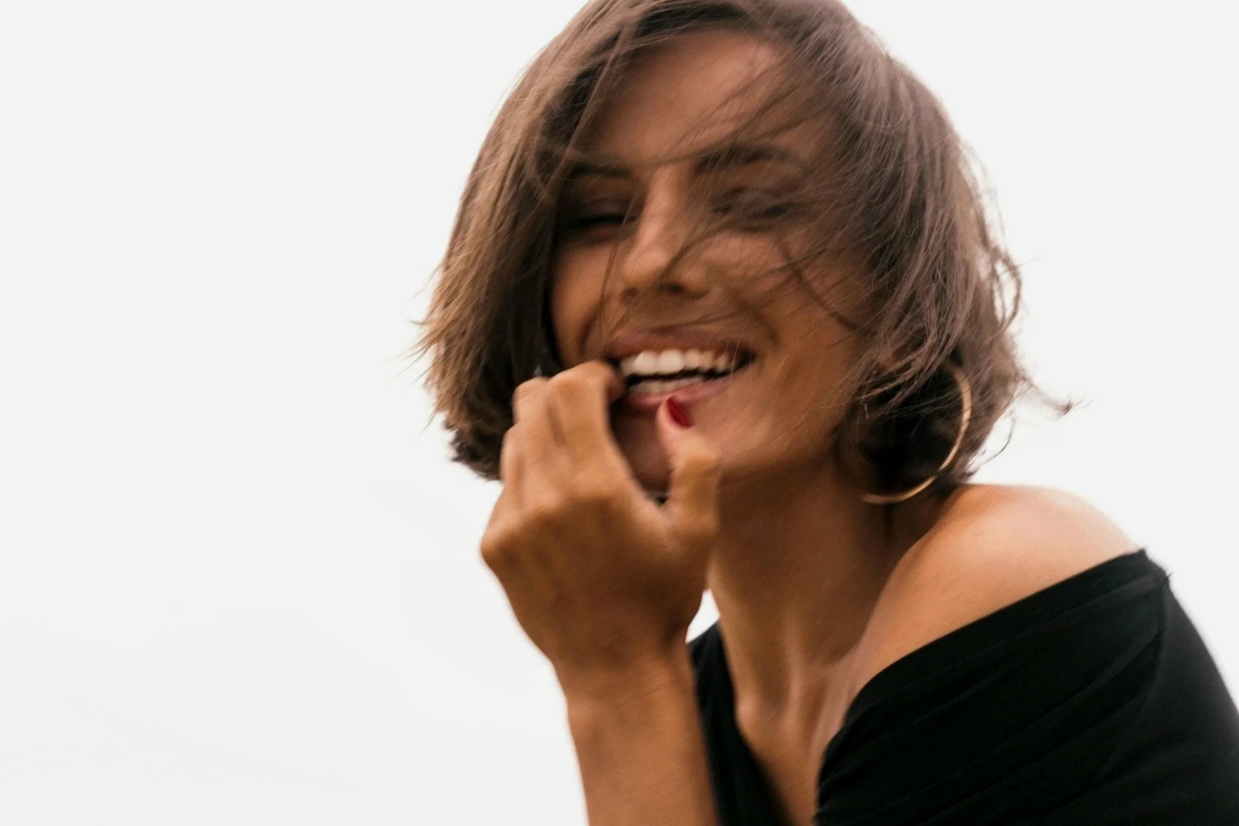 A woman with short, wavy hair smiling and touching her lips with her finger, wearing a black off-shoulder top.