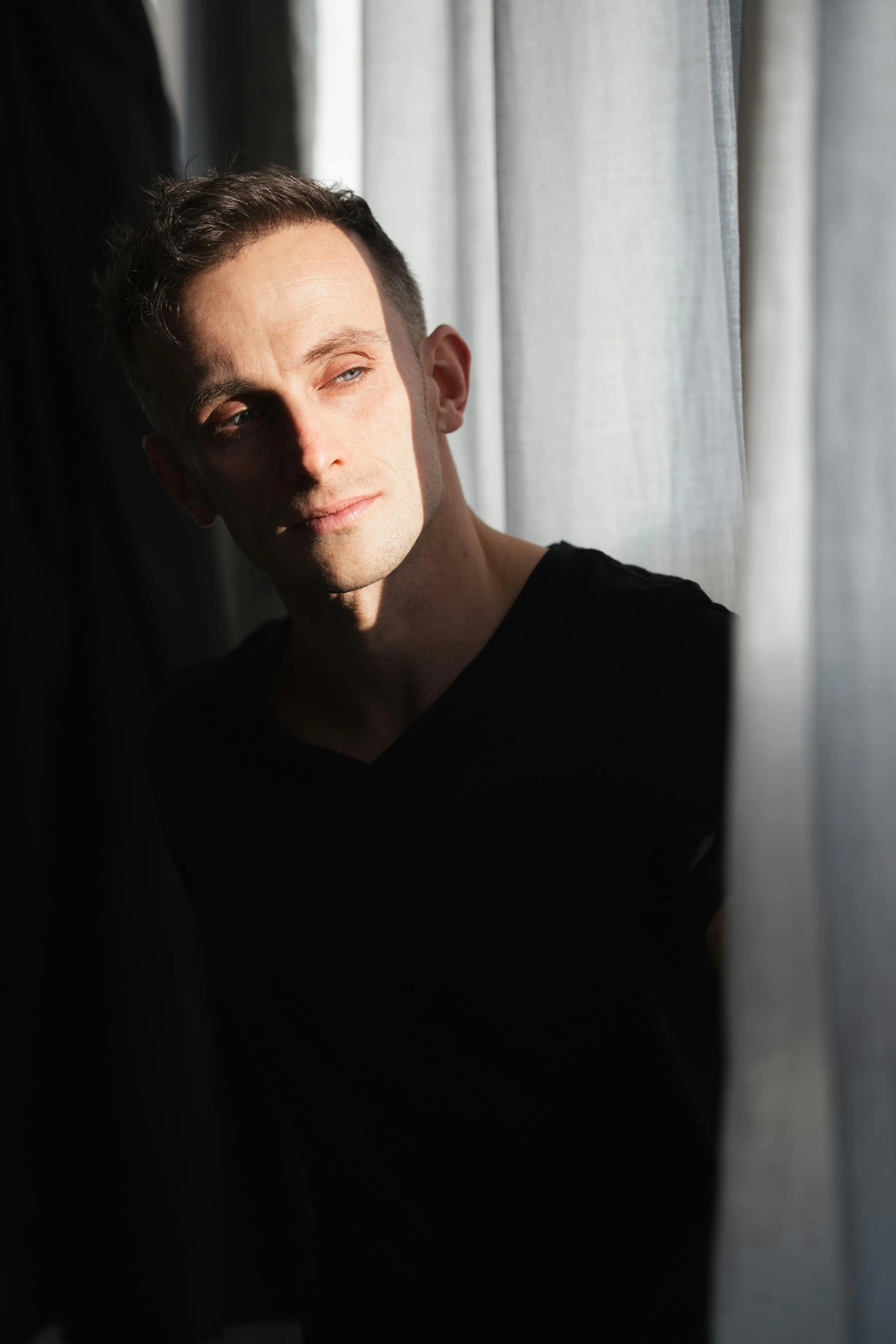 A young man with short brown hair and fair skin looking out a window, basking in sunlight, wearing a black shirt.