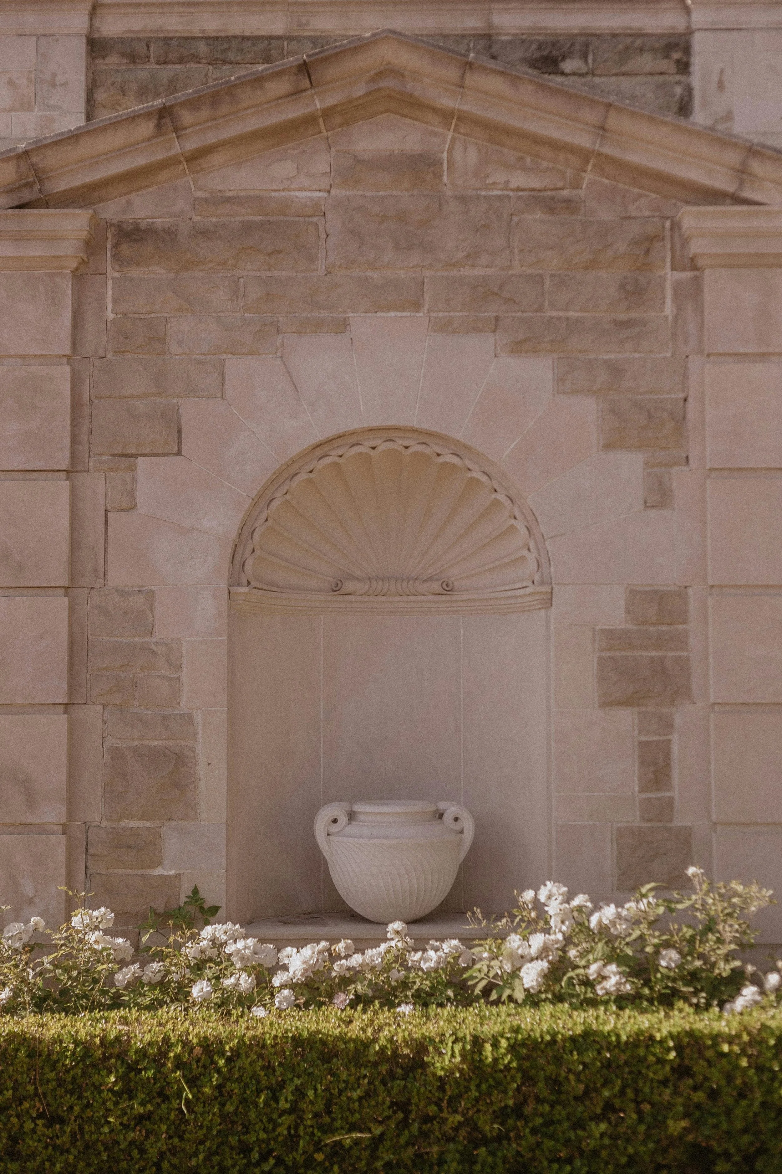 A decorative stone wall with a niche containing a classical urn sculpture, with white flowers and greenery in front.