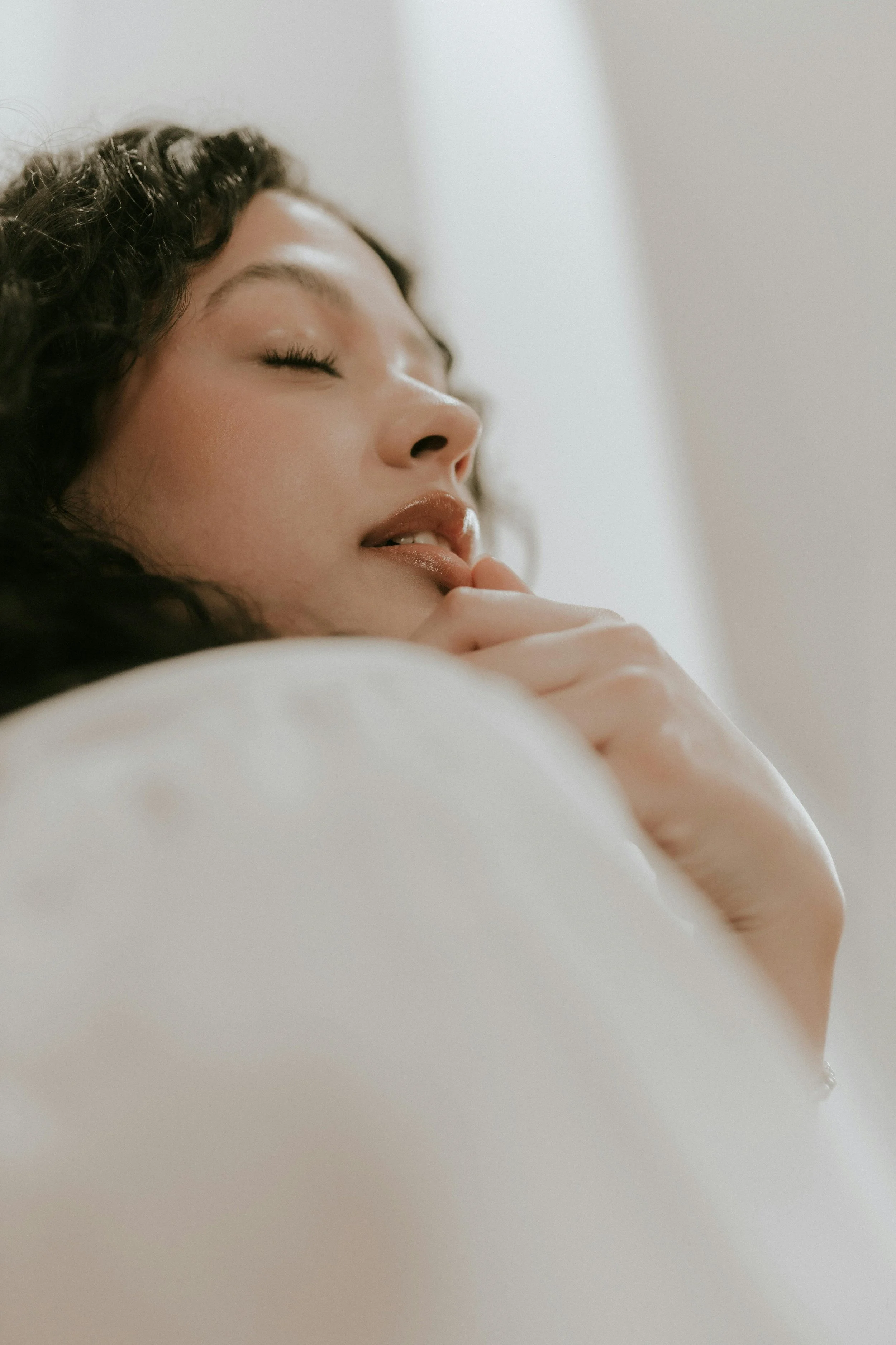 Close-up of a woman with curly hair and closed eyes, resting her chin on her hand, with a neutral and peaceful expression.