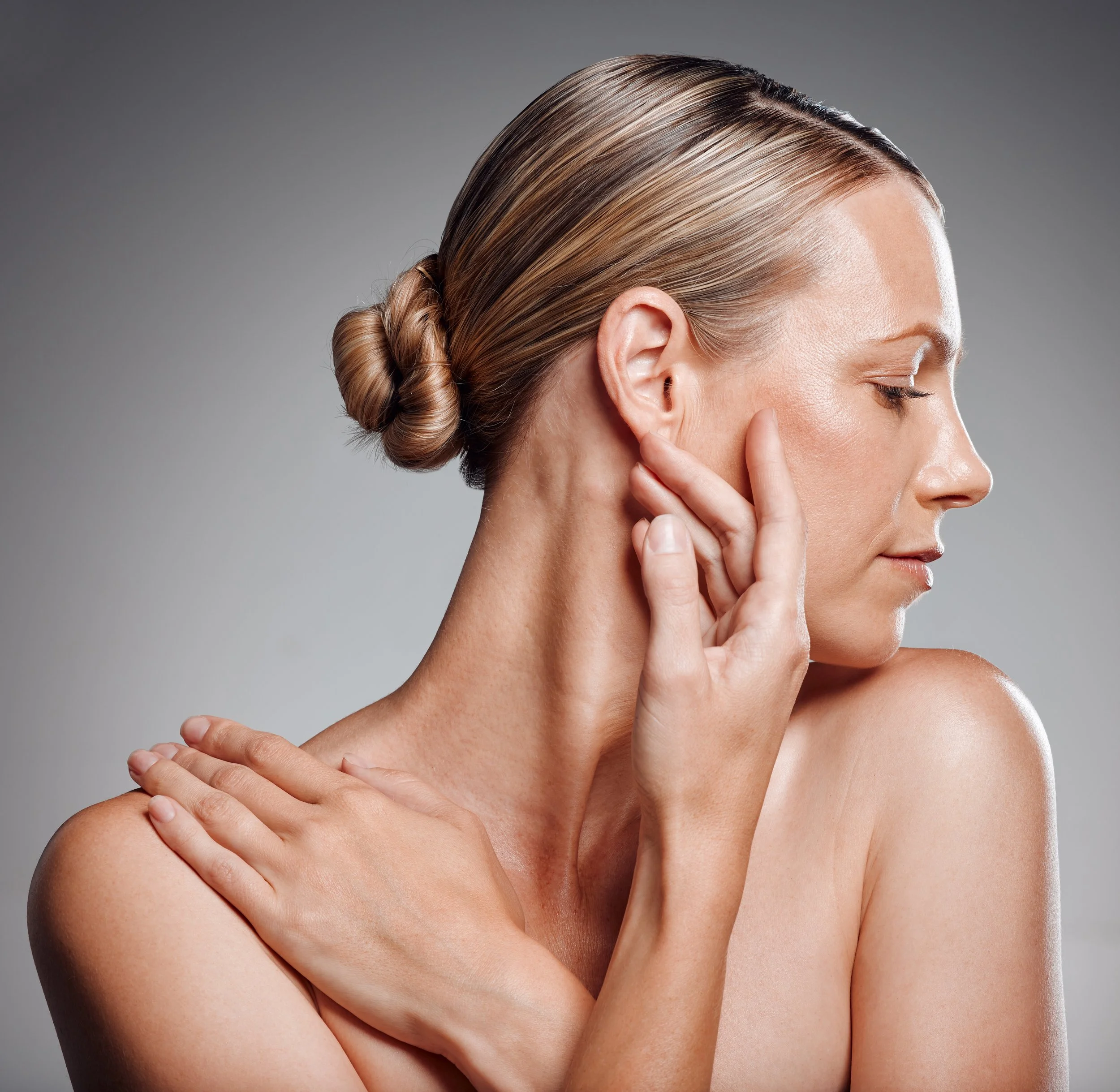 A woman with blonde hair styled in a sleek, low bun, with smooth skin and closed eyes, touching her neck and shoulder under studio lighting.