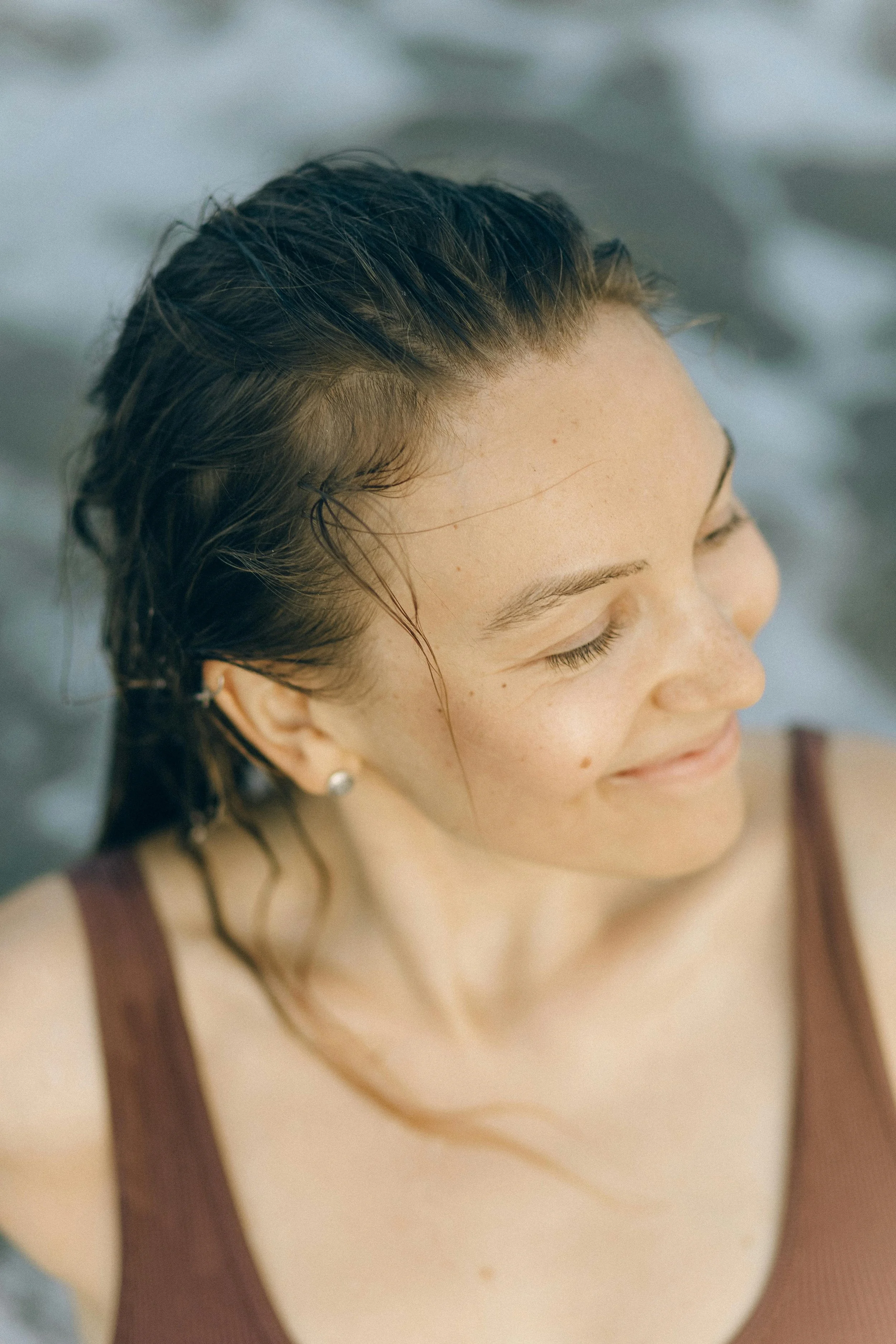 A smiling woman with wet hair and closed eyes, outdoors in a natural setting.