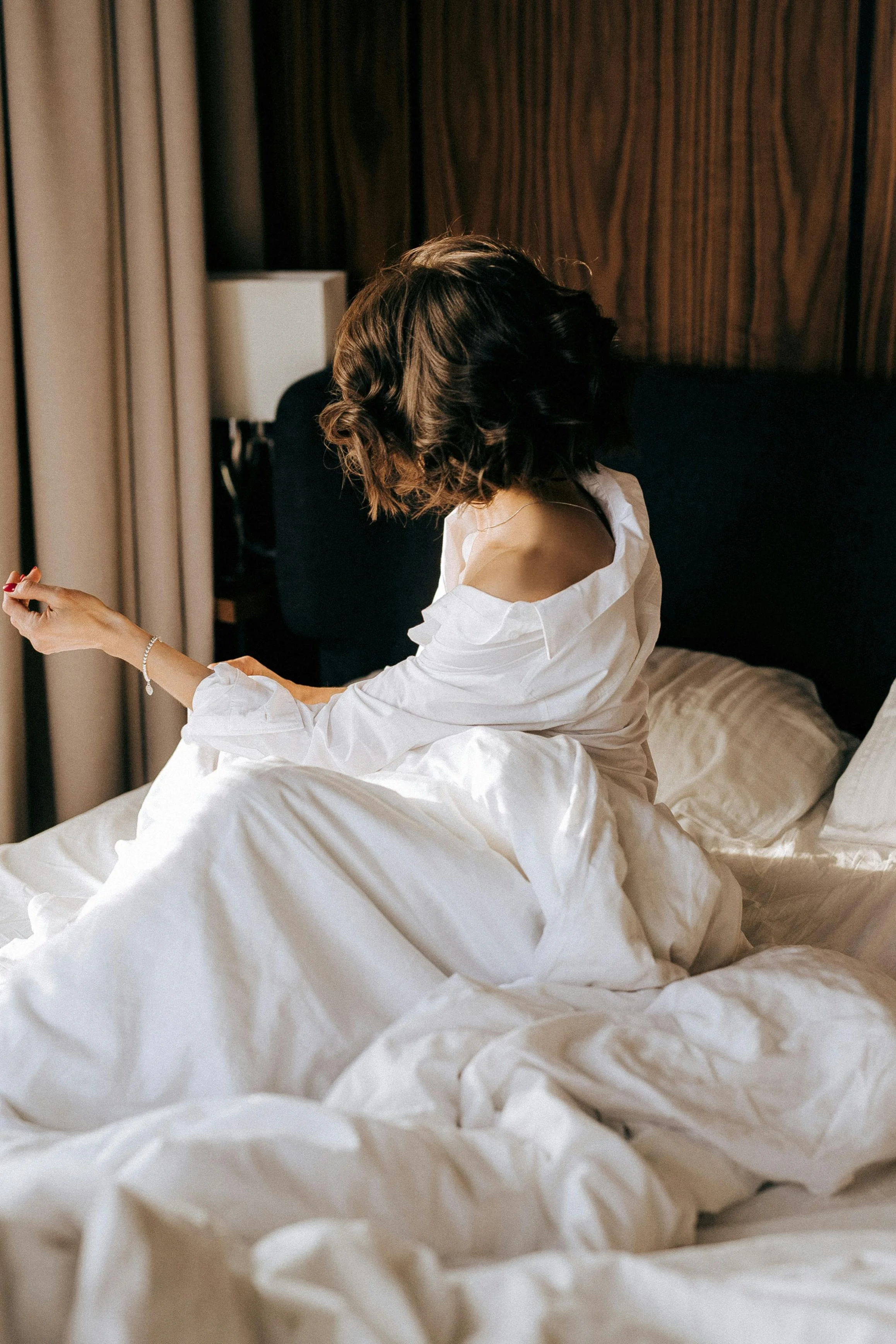 A woman with dark, wavy, shoulder-length hair sitting on a bed in a hotel room, looking away from the camera. She is wearing a white shirt that is falling off one shoulder, and she has a bracelet on her wrist. The bed has white linens, and a dark headboard is visible behind her. There is a nightstand with a lamp and curtains in the background.