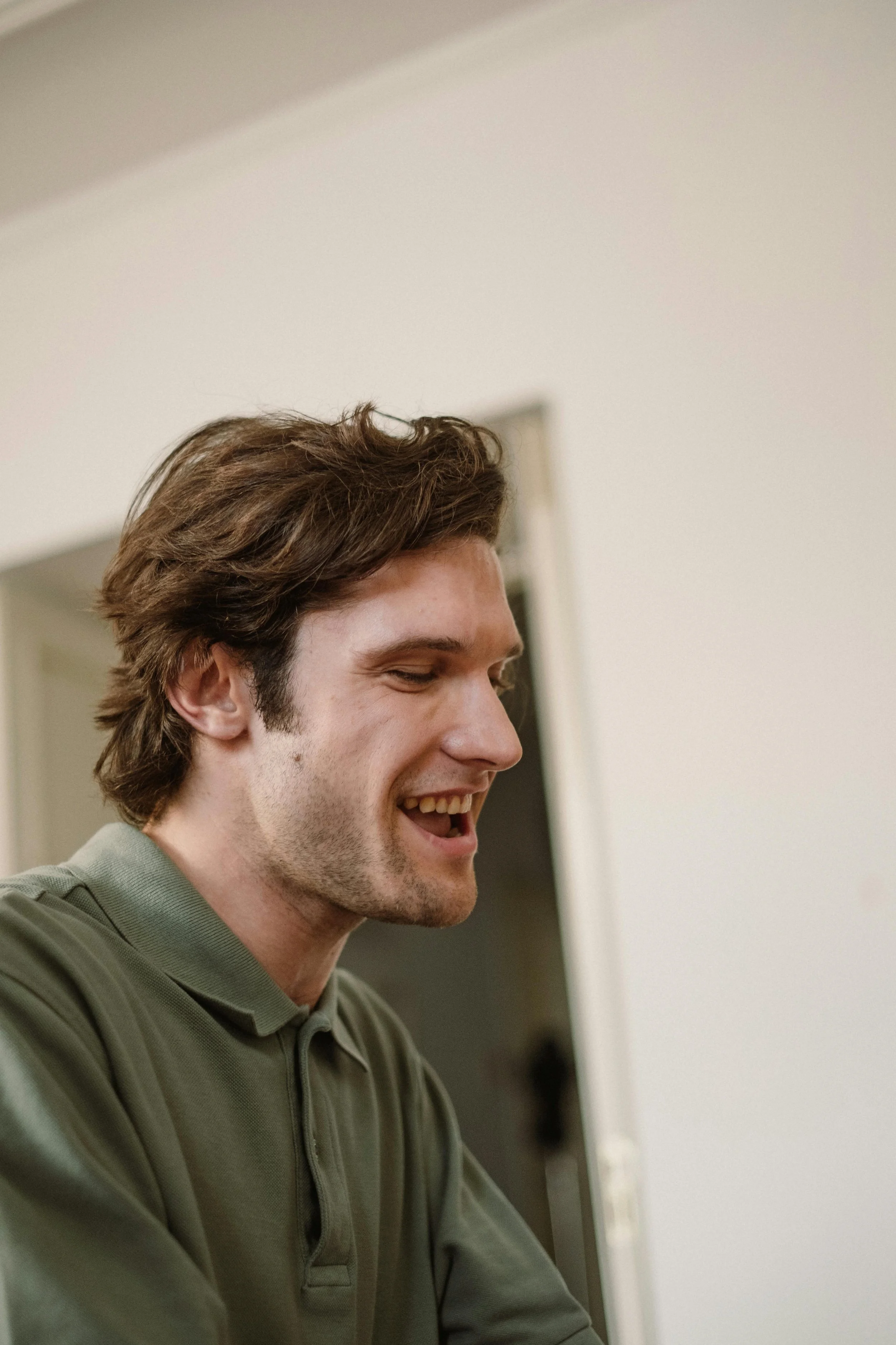 A young man with brown hair and light skin, smiling and looking down, wearing a green polo shirt, indoors.