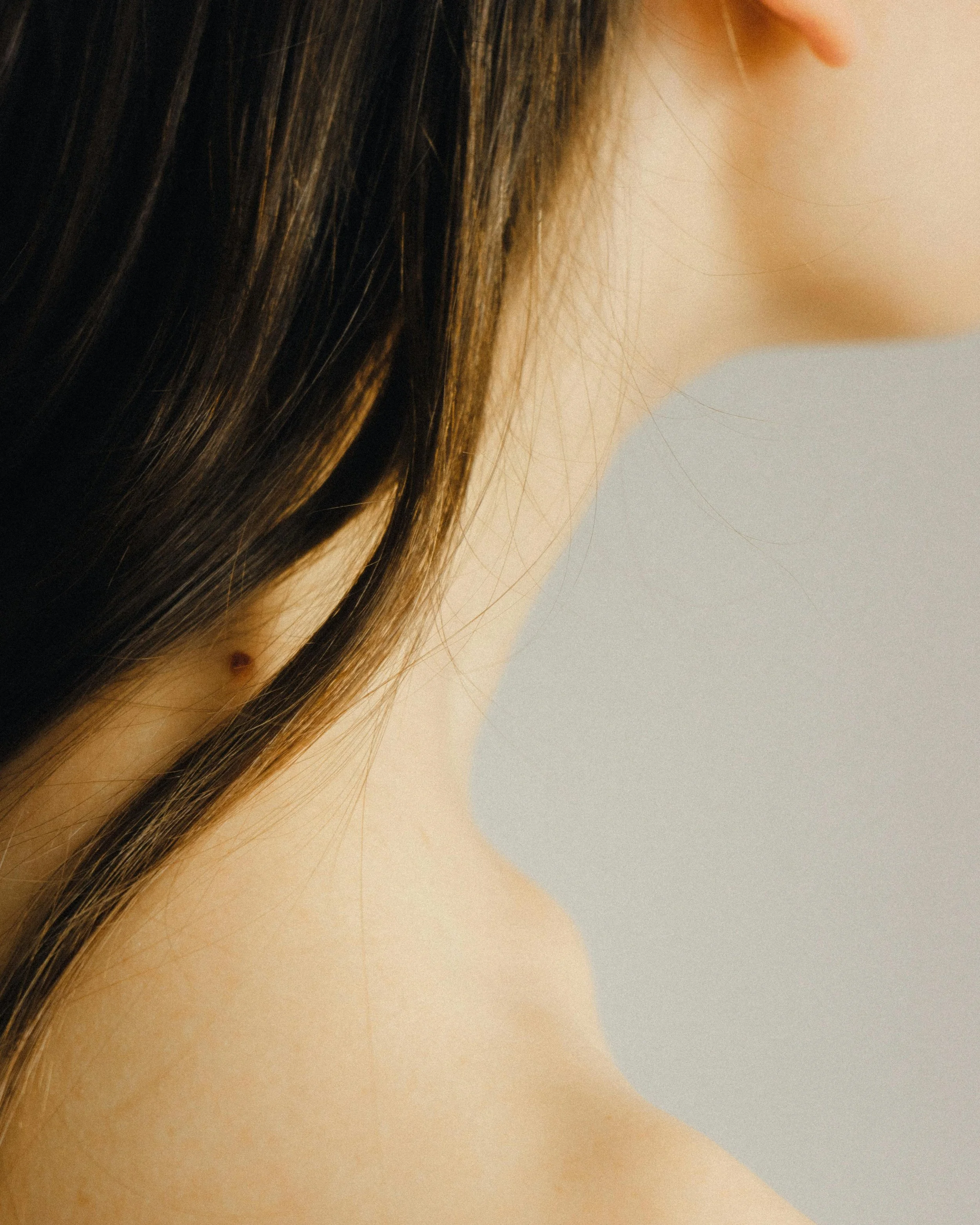 Close-up of a woman's neck and hair, with part of her face and ear visible, against a plain grey background.