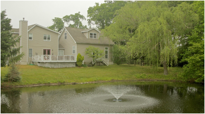 A beige two-story house with a deck, surrounded by trees and greenery, situated next to a pond with a fountain in the center.