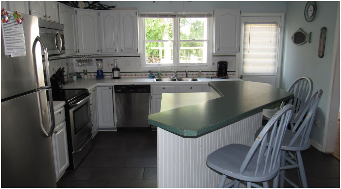 Kitchen with white cabinets, stainless steel appliances, a green countertop bar with four chairs, a window above the sink, and various small appliances on the counter.