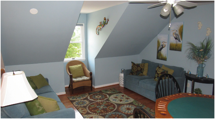 Living room with sloped ceiling, blue walls, and wooden floors, featuring two blue sofas, an armchair, a patterned rug, wall art of herons, a decorative wall gecko, and a window with natural light.