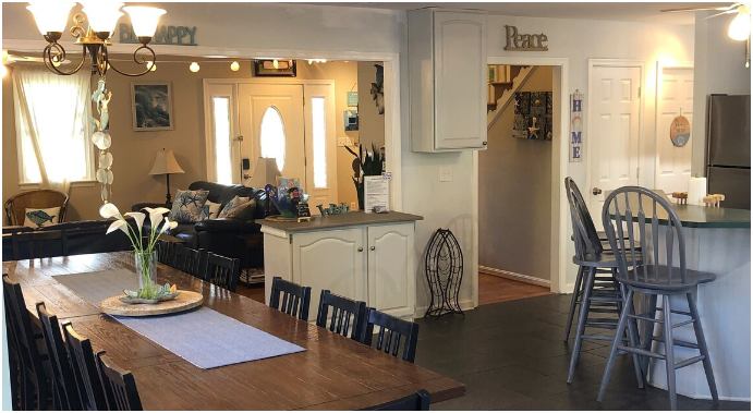 Open-concept kitchen and living room with a wooden dining table, black chairs, and a kitchen island with barstools. The living room has a couch, a side table with a lamp, and decorative signs that say 'PEACE' and 'HAPPY.' The area is warmly lit with