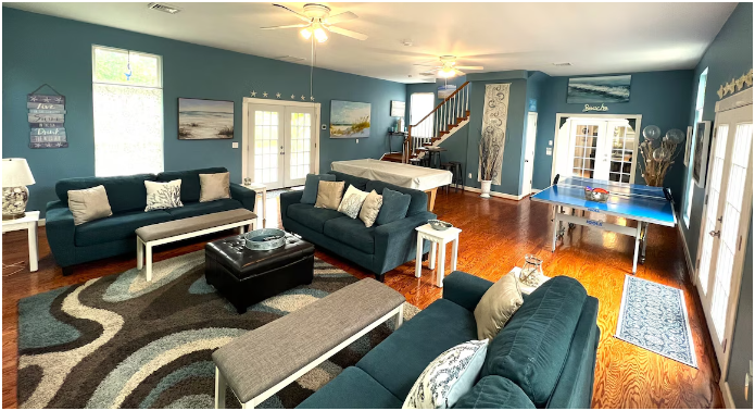 Living room with blue walls, hardwood floors, two large blue sofas, a black ottoman, a white bench, a small white side table, and a ping pong table near the sliding glass door.