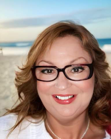 Geodie, with wavy red hair, wearing glasses and a pearl necklace, smiling at the camera with a beach and ocean in the background.
